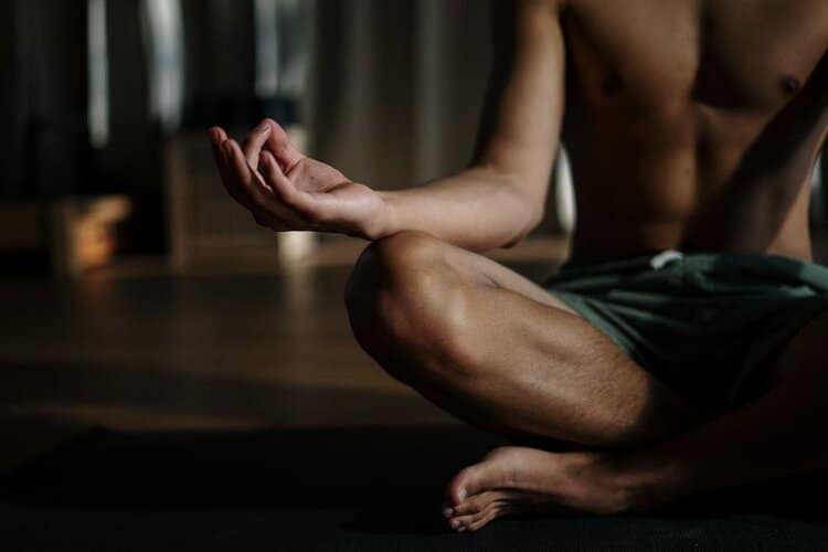 A person wearing green shorts sits cross-legged on a mat indoors, with one hand resting on their knee in a meditation pose. The background is softly lit and out of focus.