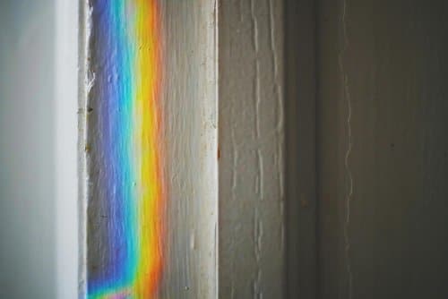 A narrow strip of rainbow light is cast onto the edge of a white painted wall, next to another plain white surface, creating a colorful effect on the textured surface.