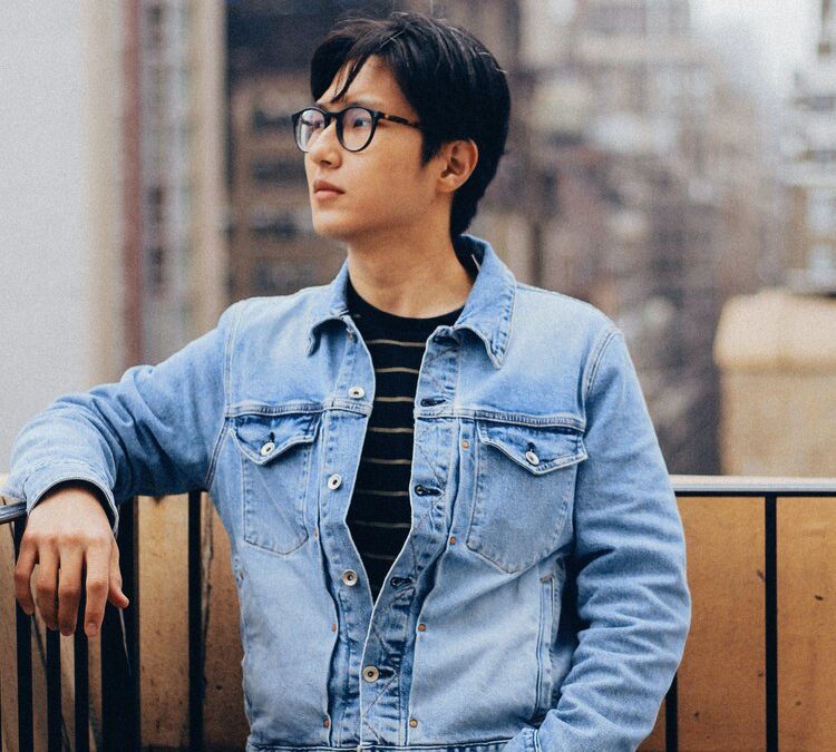 A person wearing glasses and a denim jacket stands on a balcony, resting one arm on the railing, with a cityscape of tall, blurred buildings in the background.