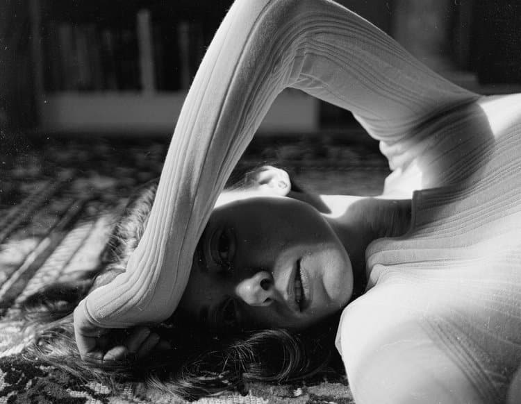 A person lies on their side on a patterned surface with one arm bent and resting across their forehead. The lighting creates strong shadows and highlights on their face and clothing. Bookshelves are blurred in the background.